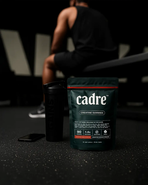 Cadre Orange Creamsicle Creatine Gummies bag on gym floor beside shaker bottle with athlete resting on bench in background, representing daily discipline and consistency.